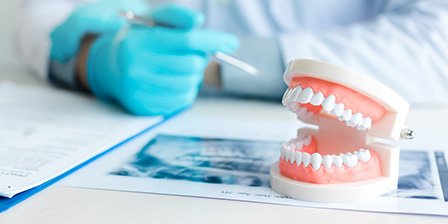 Dentist in blue gloves leaning on desk with X-ray, model teeth, and treatment plan