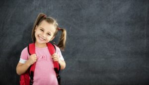 little girl wearing backpack and standing in front of a blackboard 